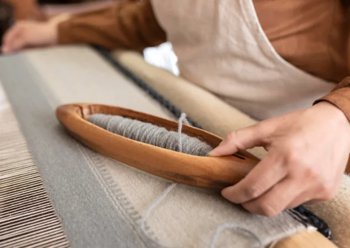 A weaver's hand using a wooden shuttle to weave grey wool