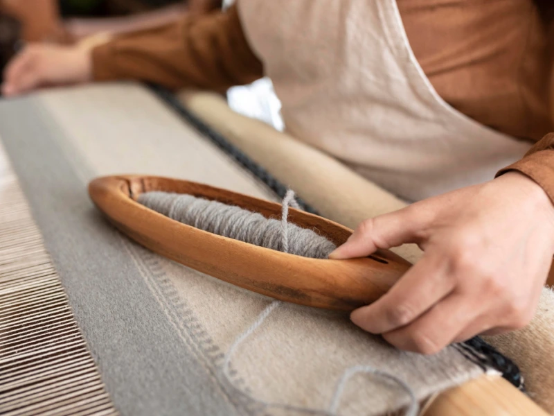 A weaver's hand using a wooden shuttle to weave grey wool